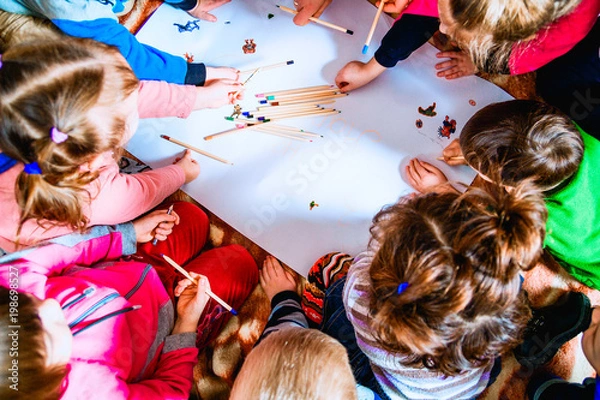 Fototapeta several small children draw on a sheet of paper with pencils. A view from above.