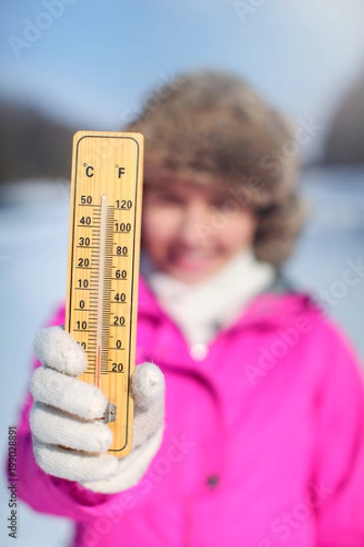 Fototapeta Young woman in pink sport jacket holding wooden thermometer outside on cold day, illustrating weather with temperature as low as -20 degrees Celsius.