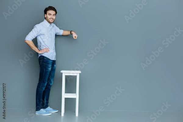 Fototapeta Useful object. Nice good looking handsome man standing near the stool and holding her hand above it while smiling to you