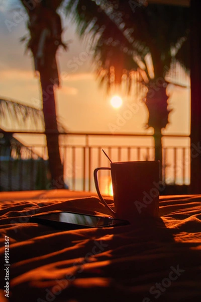 Obraz Still life. Smartphone and mug on bed amid evening sunset and silhouettes palms. Warm orange light. Concept nightfall. Copy space.