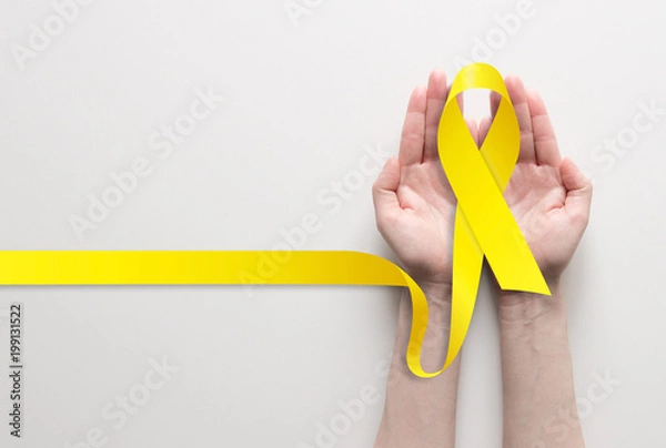 Fototapeta Dwie ręce trzymające żółtą wstążkę, symbol świadomości raka, wsparcia medycznego i profilaktyki pomocną ręką. Miejsce na tekst.