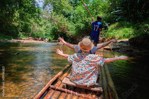 Obraz Boys (8 years old) raising one's hands floating along tropical river on bamboo raft in jungle. Concept traveling with children.