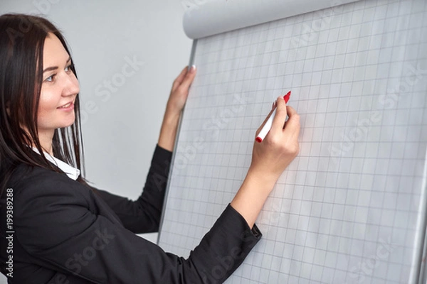Obraz Beautiful smiling young woman writing on a blank flipchart in office as she does a presentation or promotion