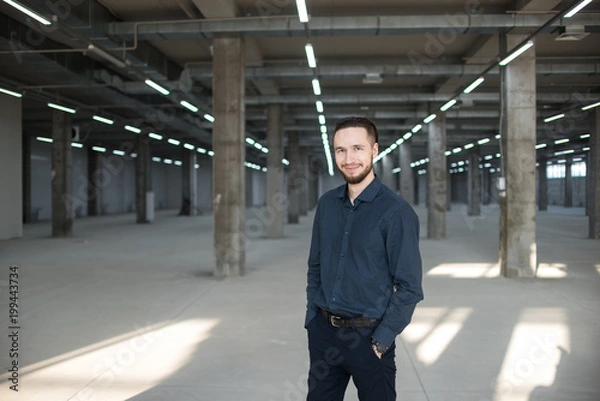 Fototapeta Young businessman on a construction site in business clothes. Beard. Hands in pockets. Dark shirt and trousers. Concrete poles.