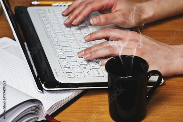 Obraz A cup of hot coffee on the table near the laptop on which is printed female fingers closeup