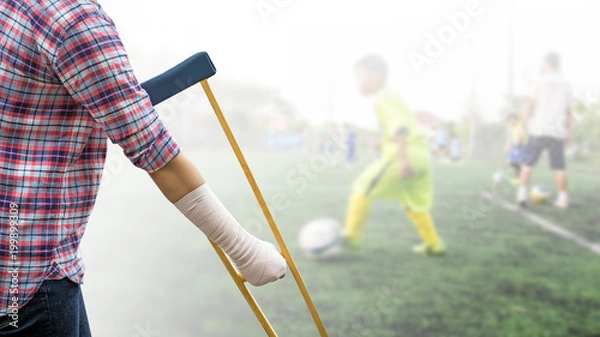 Fototapeta Injured sport player with wrist support or arm cast on hand and arm isolated on blurred kid soccer player in football field, clipping path included