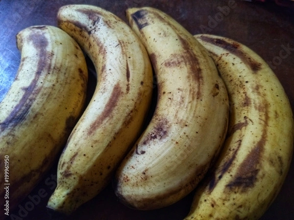 Obraz Four ripe bananas it have peels with yellow mixed a bit of brown put on the dark brown wooden table.