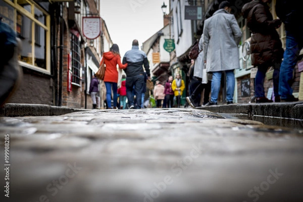 Obraz Low down shot of people and couple walking holding hands down a old traditional cobbled English street whilst browsing in shops on retail therapy day