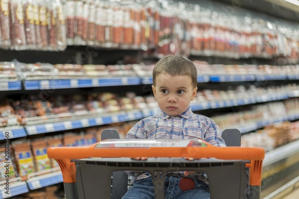 Fototapeta A cute boy dressed in a plaid shirt in a supermarket in a trolley. Against the background of shelves with meat products and sausages.