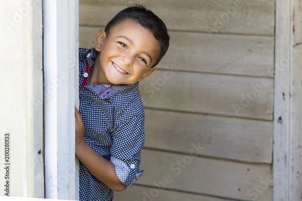 Obraz Cute little boy hiding and playing behind a door.  Outdoor portrait of a smiling little boy in blue checkered shirt.