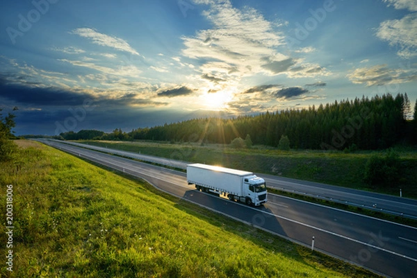 Obraz Biała ciężarówka jadąca autostradą na wsi w promieniach zachodzącego słońca z dramatycznymi chmurami