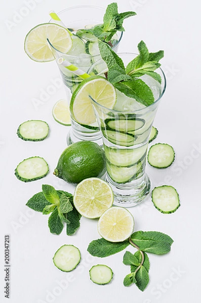 Fototapeta Fresh detox spring cocktail with mint, lime, ice, cucumber, straw on soft white wooden board as fun summer holiday background, top view, closeup.