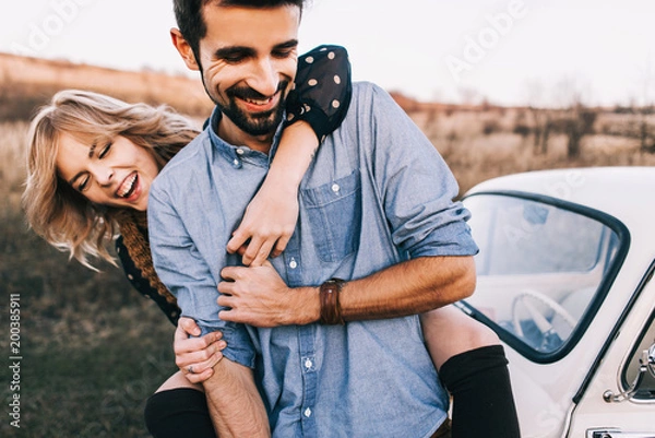 Obraz Young beautiful couple hugging each other, sitting on a small white car in beautiful evening light. Stylish guy with a beard and blond girl laughing