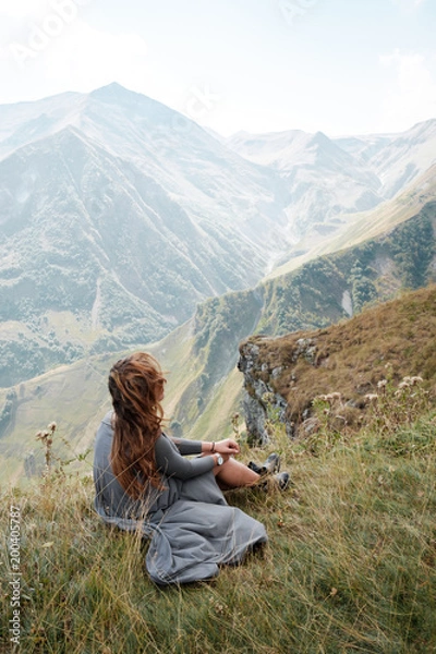 Fototapeta Beautiful young woman in a long dress is sitting on a cliff in the background of mountains