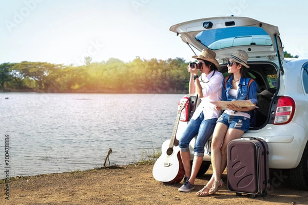 Fototapeta Couple of traveler sitting on hatchback of car and enjoying for view of nature near the lake during holiday.Young couple tourist have a guitar and baggage taking photo on vacation. 