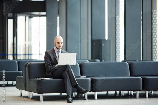 Fototapeta Young businessman with laptop sitting on black leather sofa in airport lounge and networking