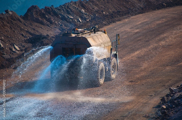 Fototapeta Heavy truck pours the road with water in the iron ore quarry. Dust removal, protection of the environment. Irrigation of the road from dust