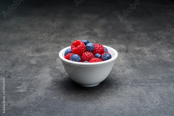 Fototapeta White ceramic bowl with berry fruit, blueberries and raspberries on dark concrete background. Close up with copy space.