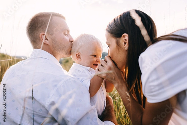 Obraz Walk beautiful young family in white clothes with a young son blond in mountainous areas with tall grass at sunset. Parents from both sides embracing son, hugging. family - this is happiness