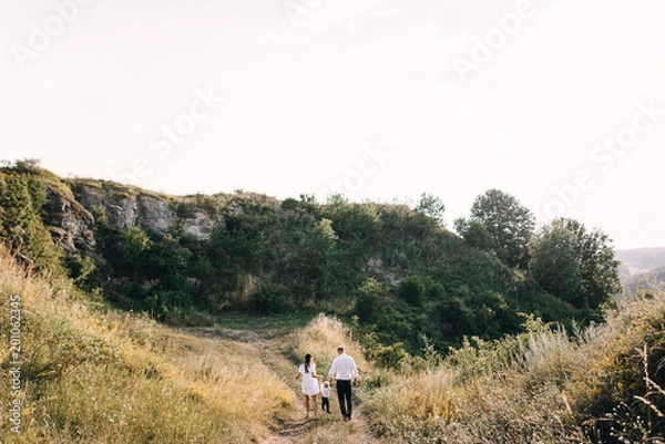 Fototapeta Walk beautiful young family in white clothes with a young son blond in mountainous areas with tall grass at sunset. Mother keeps son in her arms, hugging. family - this is happiness