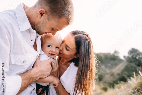 Obraz Walk of beautiful young family in white clothes with a young son blond in mountainous areas with tall grass at sunset. Dads carries his son on his shoulders. family - this is happiness