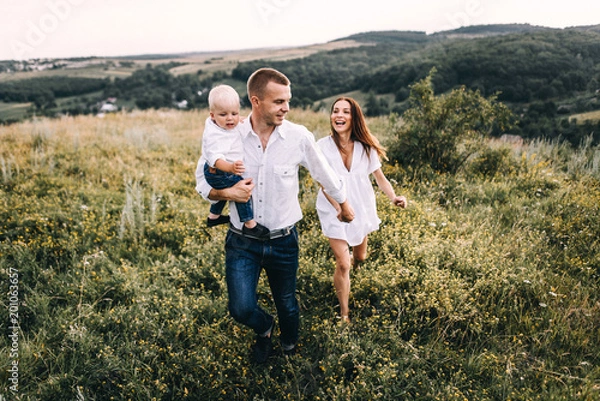 Obraz Walk beautiful young family in white clothes with a young son blond in mountainous areas with tall grass at sunset. Mother keeps son in his arms, hugging. family - this is happiness