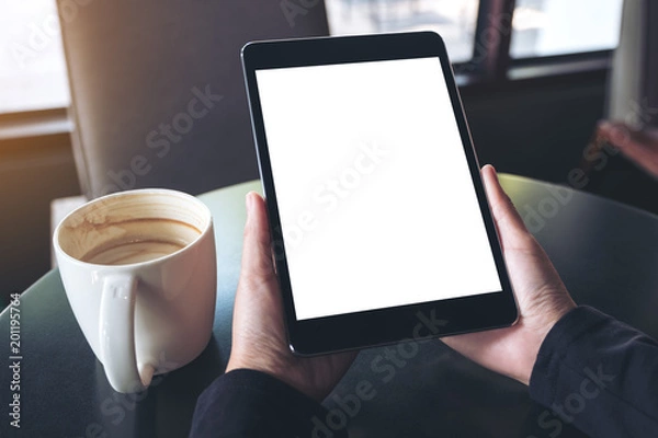 Fototapeta Mockup image of hands holding black tablet pc with blank white screen and coffee cup on table in cafe