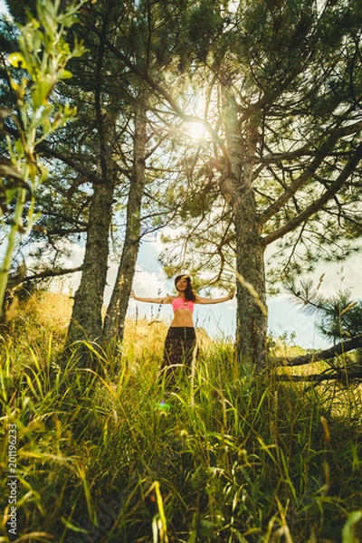 Fototapeta A young beautiful woman with an ideal figure enjoys the freshness of a summer day in a pine grove among dense grass. The sun is sparkling in the midst of the trees.