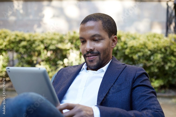 Fototapeta Smiling american african businessman in informal clothes working at sunny street on electronic touch pad,checking e-mail using Internet-enabled electronic device,calling friends via social networks