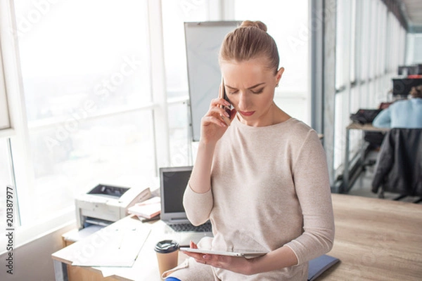 Fototapeta Business woman is looking at the tablet and talking on the phone at the same time. She has a phone meeting with client. This woman stands in a big, large room near window.