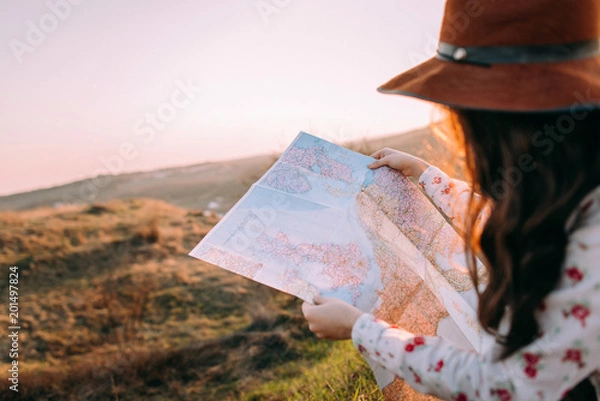 Obraz Traveler beautiful girl with a hat looking at a map holding in her hand binoculars at sunset sitting on a background of mountains. She selects a place on the map. Concept photo travel, adventure