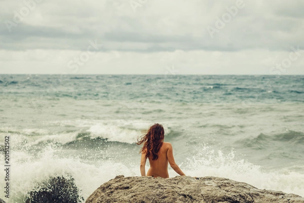 Fototapeta Naked young girl with a beautiful body stands facing the sea, back to the beach and watching the raging, mighty, strong waves during a storm in the ocean. Beach and stones. 