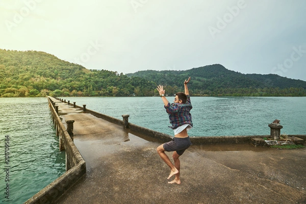 Fototapeta Traveling boy on the pier. Pretty young man jumping on the bridge. Summer lifestyle and adventure photo