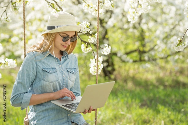 Fototapeta Portrait of a girl in a hat with glasses working on a laptop on a swing, in blooming spring gardens. The concept of freelancing