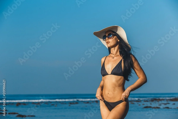 Obraz Beach vacation. Hot beautiful woman in sunhat and bikini standing with her arms raised to her head enjoying looking view of beach ocean on hot summer day. Photo from Hapuna beach, Big Island, Hawaii.