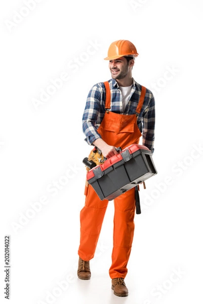 Fototapeta Smiling builder in orange overall and helmet carrying tool box isolated on white