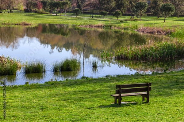 Obraz Garden bench overlooking the lake or pond of Parque da Devesa Urban Park in Vila Nova de Famalicao, Portugal. Built near the center of the city. View of the green grass lawns, and lake.