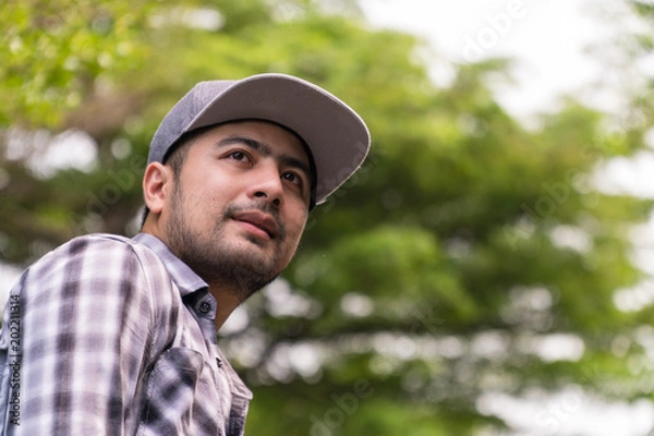 Fototapeta Portrait Handsome man : Good looking young man. He has a beard and wears a hat. He has an interesting smile. Looking for someone. Behind him, full of green trees. Copy space.
