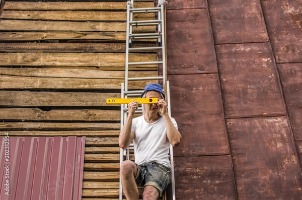 Fototapeta A man sits on the roof on a ladder with a building level