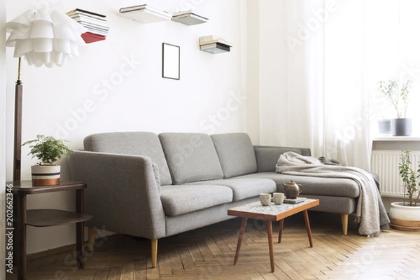 Fototapeta Design scandinavian interior of living room with small stylish table and sofa. White walls with shelves from books and mock up frame, plants on the windowsill. Brown wooden parquet.