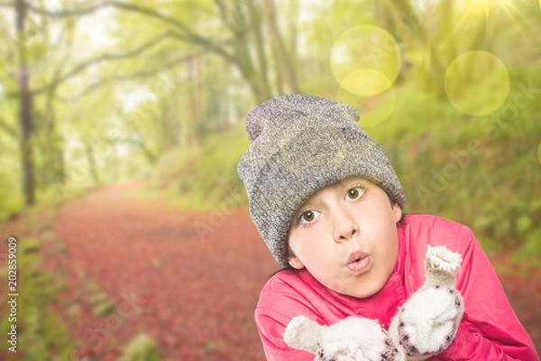 Fototapeta Wrapped up little girl blowing over hands against peaceful autumn scene in forest