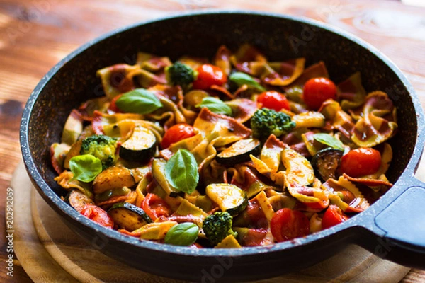 Fototapeta Włoski makaron farfalle w sosie pomidorowym i różnego rodzaju warzywami na drewnianym tle, wolne miejsce na tekst.