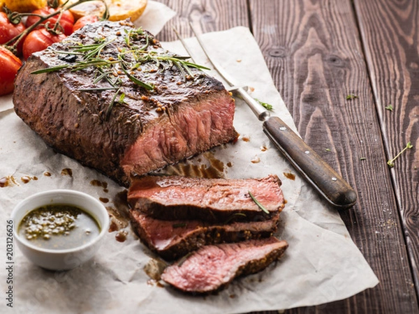 Obraz Sliced Roast beef on white paper on wooden table with grilled vegetables.