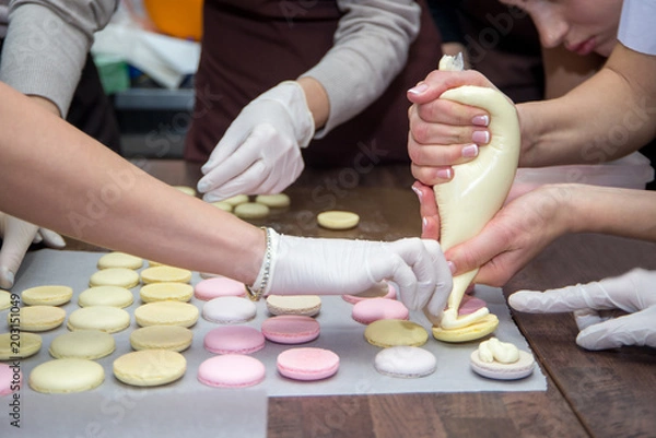 Fototapeta cooking, food and baking concept - chef with confectionery bag squeezing cream filling to macarons shells at pastry shop