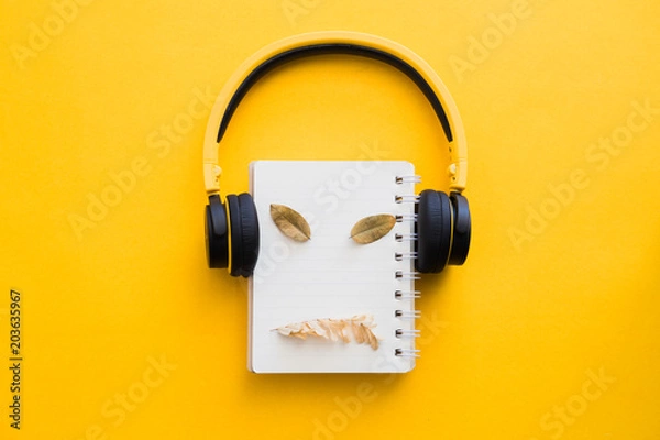 Fototapeta Yellow headphone with blank notebook and dry leaves on yellow paper background