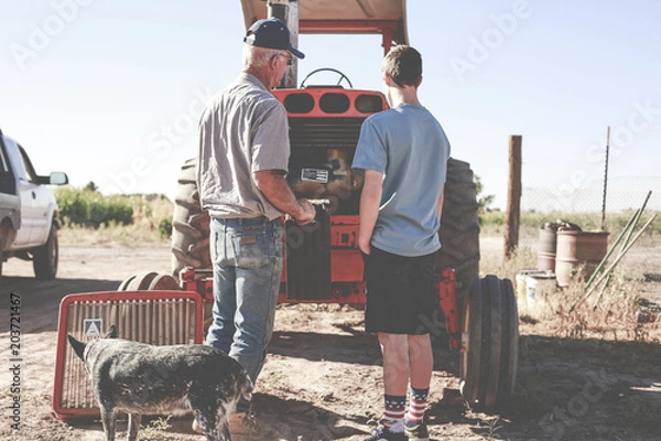 Fototapeta Man teaching boy how to fix a tractor on their farm
