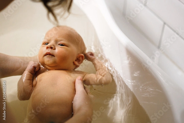 Fototapeta The newborn swims in the bathroom in the arms of the mother. The first bathing of the baby in the bathroom.