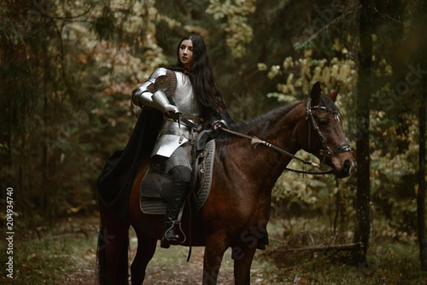 Fototapeta A beautiful warrior girl with a sword wearing chainmail and armor riding a horse in a mysterious forest.