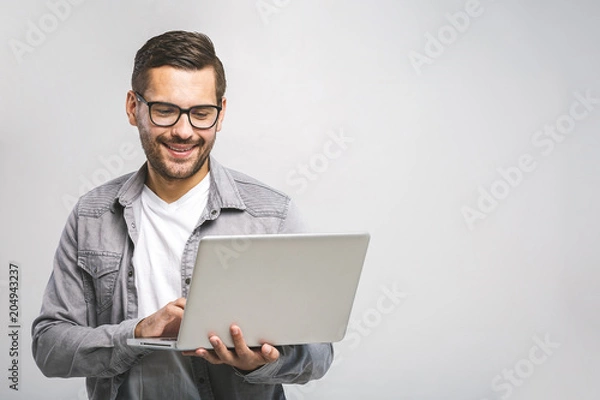 Fototapeta Confident business expert. Confident young handsome man in shirt holding laptop and smiling while standing against white background