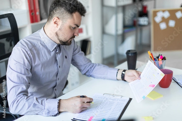 Fototapeta A young man in the office sits at a table, holds a marker in his hand and works with documents.